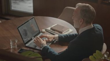 Kafkasyalı beyaz saçlı, şık formalı iş adamının göğüsleri. Restorandaki laptopta çalışırken sunumdaki iş istatistiklerini analiz ediyor.