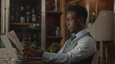 Genç, kendine güvenen Afro-Amerikan iş adamının bel hizası mali raporları analiz ediyor ve modern restoranda uzaktan çalışan bir dizüstü bilgisayar kullanıyor.