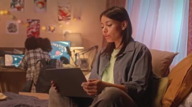 Küçük kızıyla evde otururken dizüstü bilgisayarında sohbet eden genç bir kadının orta boy görüntüsü.