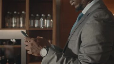 Zarif gri takım elbiseli genç Afrikalı Amerikalı iş adamının modern restoranda barla dururken akıllı telefondan mesajlaşmasını yukarı kaldır.