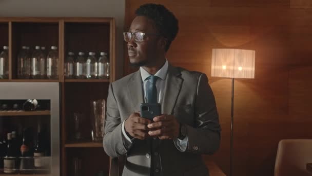 暖かいライトの豪華なレストランで立っているスマートフォンを使用して灰色のスーツのエレガントな若いアフリカ系アメリカ人のビジネスマンの中型ショット — ストック動画