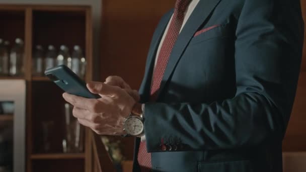 エレガントなスーツと高価な腕時計を身に着けている未知の成功したビジネスマンが屋内でスマートフォンでスクロール — ストック動画