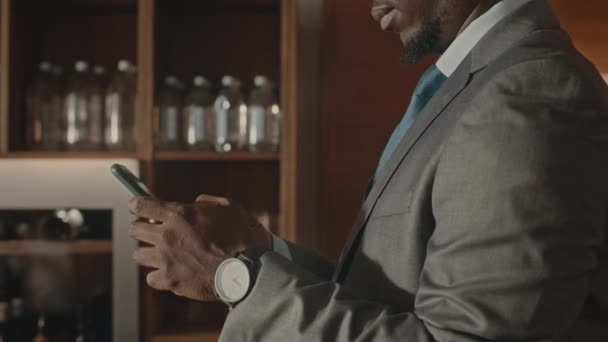 成功した若いアフリカ系アメリカ人のビジネスマンのショットは バーとモダンなレストランに立っている間 スマートフォンでエレガントな灰色のスーツのテキストで — ストック動画