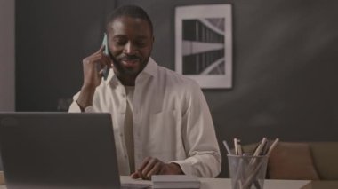 Ofis masasında dizüstü bilgisayara karşı otururken cep telefonuyla konuşan genç Afrikalı Amerikalı iş adamının belini kaldırın.
