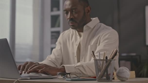 最小限のオフィスで机に座っている間 ラップトップコンピュータにタイピングする集中的な若い黒人男性起業家のウエスト — ストック動画