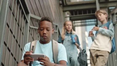 Beyaz sınıf arkadaşları arka planda sohbet ederken Afrikalı Amerikalı öğrencilerin okuldan sonra merdivenlerde oturup akıllı telefonlarıyla oyun oynamaları düşük bir açı.