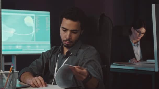 集中した中東の男性サイバーセキュリティワーカーが夜のオフィスデスクでコンピュータで座っている間に書類ワークをしています — ストック動画