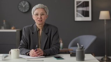 Orta yaşlı, zarif ceketli ve gözlüklü Asyalı iş kadınının bel altı görüntüsü modern grafit duvarlı ofiste masasında oturuyor ve kameraya konuşuyor.
