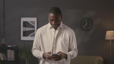 Genç Afrikalı Amerikalı iş adamının orta boy portresi akıllı telefondan bakınıyor ve minimalist ofiste kameraya poz veriyor.