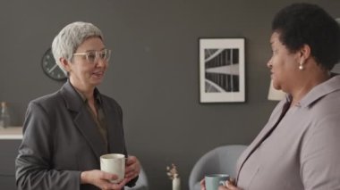 Orta yaşlı, orta yaşlı iki kadın meslektaşın iş yerinde minimalist grafit duvarlı ofiste sohbet ederken orta boy bir fotoğraf.