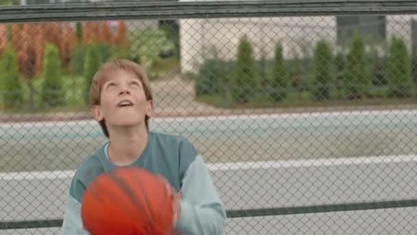 夏の屋外裁判所に立って バスケットボールと笑顔で遊ぶクールなコーカサス小児のミディアムスローモの肖像画 — ストック動画