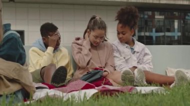 Bir grup çok ırklı anaokulu öğrencisi yazın açık yeşil çimlerde birlikte oturuyorlar. Akıllı telefon kullanıyorlar ve sohbet ediyorlar.