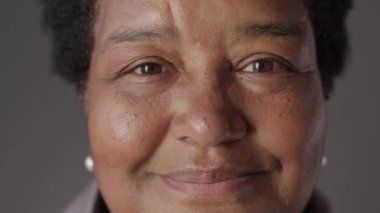 Gri stüdyo arka planında kameraya nazikçe bakan yaşlı siyahi kadının yakın plan portresi.