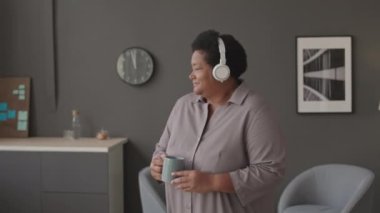 Orta boy olgun, iri yarı, elinde bir kupa çayla minimalist ofiste kablosuz kulaklıkla müzik eşliğinde dans eden zenci bir kadın.
