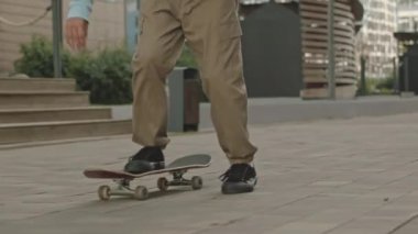 Yazın sokakta kaykay süren, bol deve pantolonlu ve spor ayakkabılı, tanınmayan bir gencin görüntüsü.