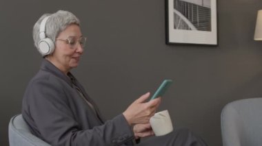Orta yaşlı Asyalı iş kadınının kablosuz kulaklıkla müzik dinlediği, akıllı telefonla kaydığı ve işyerinde ofis molası verip kahve içtiği orta boy bir fotoğraf.