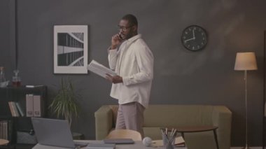 Orta boy Afrikalı Amerikalı iş adamı elinde mali kağıt raporuyla iş ortağıyla cep telefonuyla konuşurken, merkez büroda ileri geri yürürken.