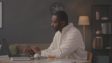 Dizüstü bilgisayarda çalışan genç siyahi iş adamının belinde bir bardak viski ve kağıt dökümanlarıyla ofis masasında oturuyor.