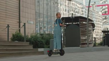 Güneşli bir günde kaldırım boyunca elektrikli scooter süren, kot takım elbiseli, beyaz tenli, genç bir kız.