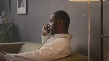 Yakışıklı siyahi girişimcinin belden yukarısı minimalist ofiste yumuşak koltukta oturmuş telefonla konuşuyor.
