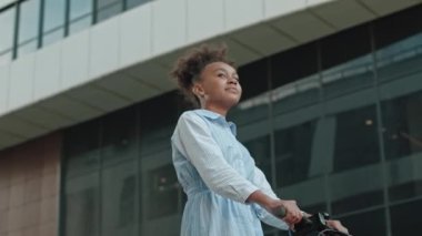 Düşük açılı Afro-Amerikalı, gökyüzü mavisi yaz elbiseli kız şehirde elektrikli scooter sürerken etrafa bakıp gülümsüyor.