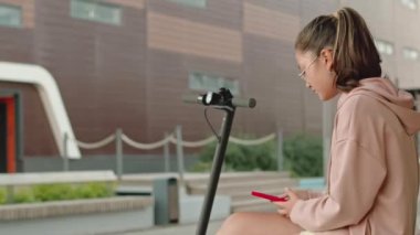Kafkasyalı genç bir kızın akıllı telefonuyla arkadaşlarıyla mesajlaşırken ahşap bankta otururken sonra da elektrikli scooterın üzerinde dikilirken ve uzaklaştığı orta boy bir fotoğraf.