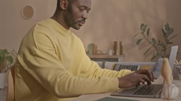 在明亮的客厅里 穿着黄色运动衫的黑人年轻人站在笔记本电脑旁边 看着摄像机 在家里自由职业工作者的面前 举着他的肖像 — 图库视频影像