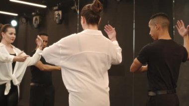 Beyaz gömlekli ve kırmızı dudaklı genç beyaz kadın aynanın önünde dikiliyordu. Çift ırklı erkek öğretmen geniş stüdyoda bireysel sınıfta Latina dansı öğreniyordu.