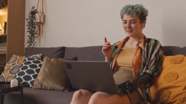 Orta boy gülümseyen, protez kollu, beyaz bir kadının, sıcak bir ışıkla sıcak bir oturma odasında kanepede otururken arkadaşıyla bilgisayarla görüntülü konuşma görüntüsü.