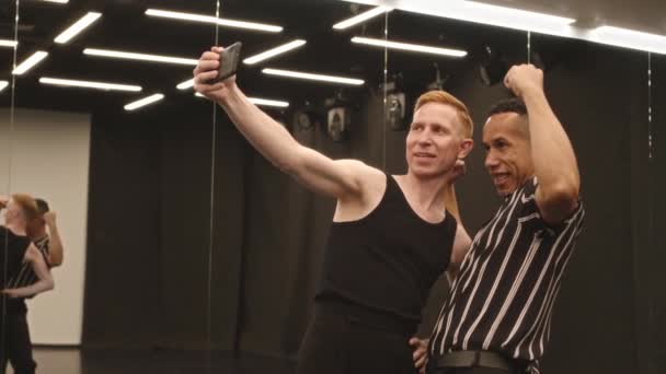 広々としたボールルームでスマートフォンカメラで面白いセルフィーの肖像画を撮る2人の多様な男性のボールルームダンサーの中央スローモ — ストック動画