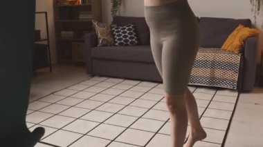 Spor kıyafetleri içinde, evde antrenman yaparken yoga minderini salonun zeminine seren, tanınmayan genç ve aktif bir kadının görüntüsü.