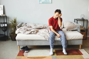 Genç adam yatağında oturuyor ve sabahları evde uyanıyor.