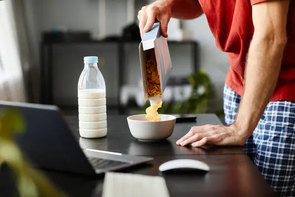 ボウルにトウモロコシフレークを入れて ラップトップ付きのテーブルで朝食のためにミルクでそれらを作る若者のクローズアップ — ストック写真