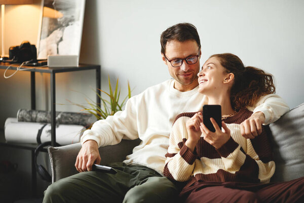 Young couple resting on sofa, using mobile phone and talking during leisure time