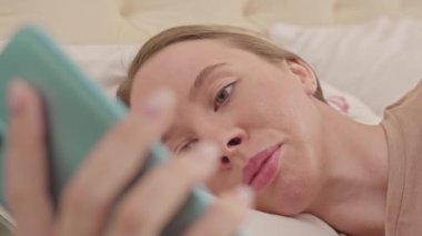Genç, güzel, beyaz bir kadının akıllı telefonuyla göz gezdirirken sabahları yatakta rahat bir şekilde yatarken orta ölçekte çekilmiş fotoğrafı.