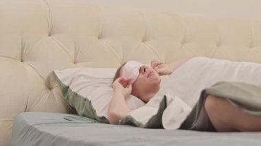Güzel beyaz kadın, sabahları rahat bir yatakta uyandıktan sonra uyku maskesini çıkarıyor ve akıllı telefonunu alıyor.