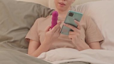 Yatakta elinde mor vibratör ve akıllı telefonuyla oturan genç bir kadın kendi kendine oyun oynayacak.