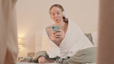 Genç beyaz kadın yatakta omuzlarında battaniyeyle oturuyor, kablosuz kulaklıkla müzik dinliyor ve akıllı telefon kullanıyor, sabahını evde yalnız geçiriyor.
