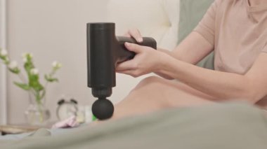 Modern bir dairede rahat bir yatakta otururken bacak kaslarında masaj tabancası kullanan tanınmayan genç bir kadının görüntüsü.