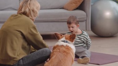 Neşeli 6 yaşında, Down sendromlu bir çocuk. Parlak oturma odasında halıya oturmuş annesiyle ve şirin Corgi köpeğiyle oynuyor.