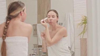 Duştan sonra beyaz havlu giyen beyaz bir kadın banyoda aynanın önünde cilt bakımı yaparken gözlerinin altına göz sıvısı serumu sürüyor.