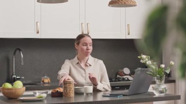 Orta boy beyaz bir kadın mutfak masasında dizüstü bilgisayarın başında oturup müsli kahvaltısı hazırlıyor.