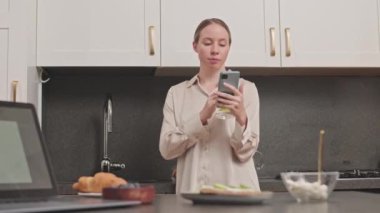 Orta boy beyaz bir kadın limonlu su içerken ve akıllı telefon kullanırken modern mutfakta ipek pijama gömleği giyerken.