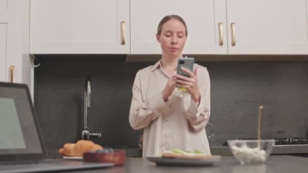 中镜头的年轻白种人美女喝着一杯柠檬水 穿着丝绸睡衣站在现代厨房里用智能手机 — 图库视频影像
