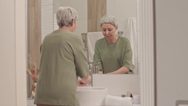 在简约的浴室里 一位年长的亚洲女人站在镜子前 用白色毛巾洗脸和擦拭脸 — 图库视频影像