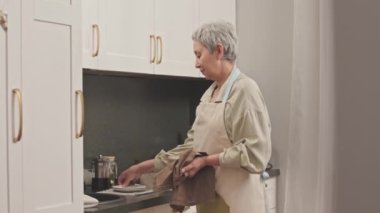 Orta boy, gri saçlı, yaşlı, Asyalı bir kadının tabakları havluyla kurutup en alt raftaki beyaz mutfak dolabına koyması.