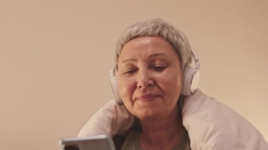 Düşük açılı gülümseyen yaşlı Asyalı kadın omuzunda yumuşak battaniyeyle gece yatağında akıllı telefonuyla geziniyor.