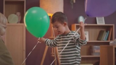Down sendromlu, mutlu, küçük beyaz bir çocuğun, şefkatli annesinin hediye ettiği renkli balonları elinde tuttuğu bir fotoğraf.