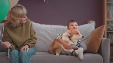 Neşeli Kafkasyalı küçük çocuk Down sendromlu sevimli Corgi köpeğini kucaklıyor parlak sıcak oturma odasında kanepede birlikte oturuyor. Çocuklar ona balon veriyor.