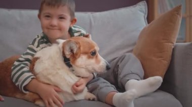 Morali bozuk, 6 yaşında beyaz bir çocuğun tatlı Corgi köpeğini nazikçe kucaklaması, gündüz vakti oturma odasında kanepede birlikte oturması.